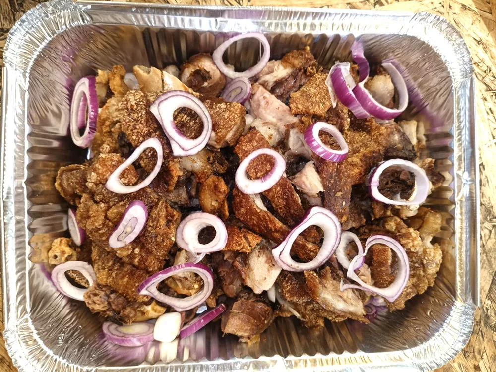 An aluminum tray filled with a serving of Tita Ping's Crispy Pata topped with onion slices.